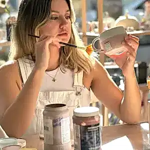 Atölye Dally seramik atölyesinde, genç bir kadın el yapımı seramik kupayı özenle fırça ile boyuyor, sanatsal üretim sürecini sergiliyor.